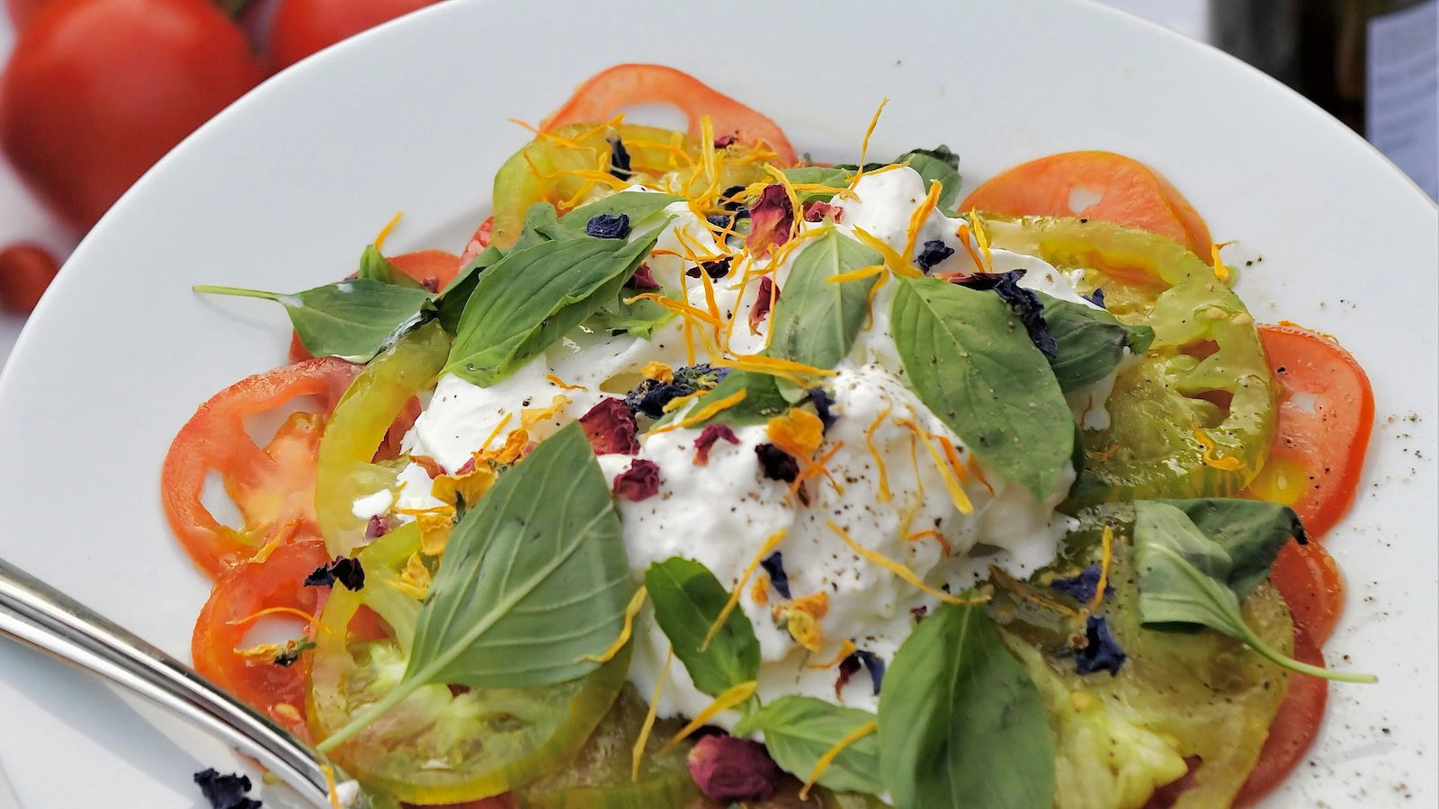 A plate of caprese salad with a fork and a bottle of wine in the background.