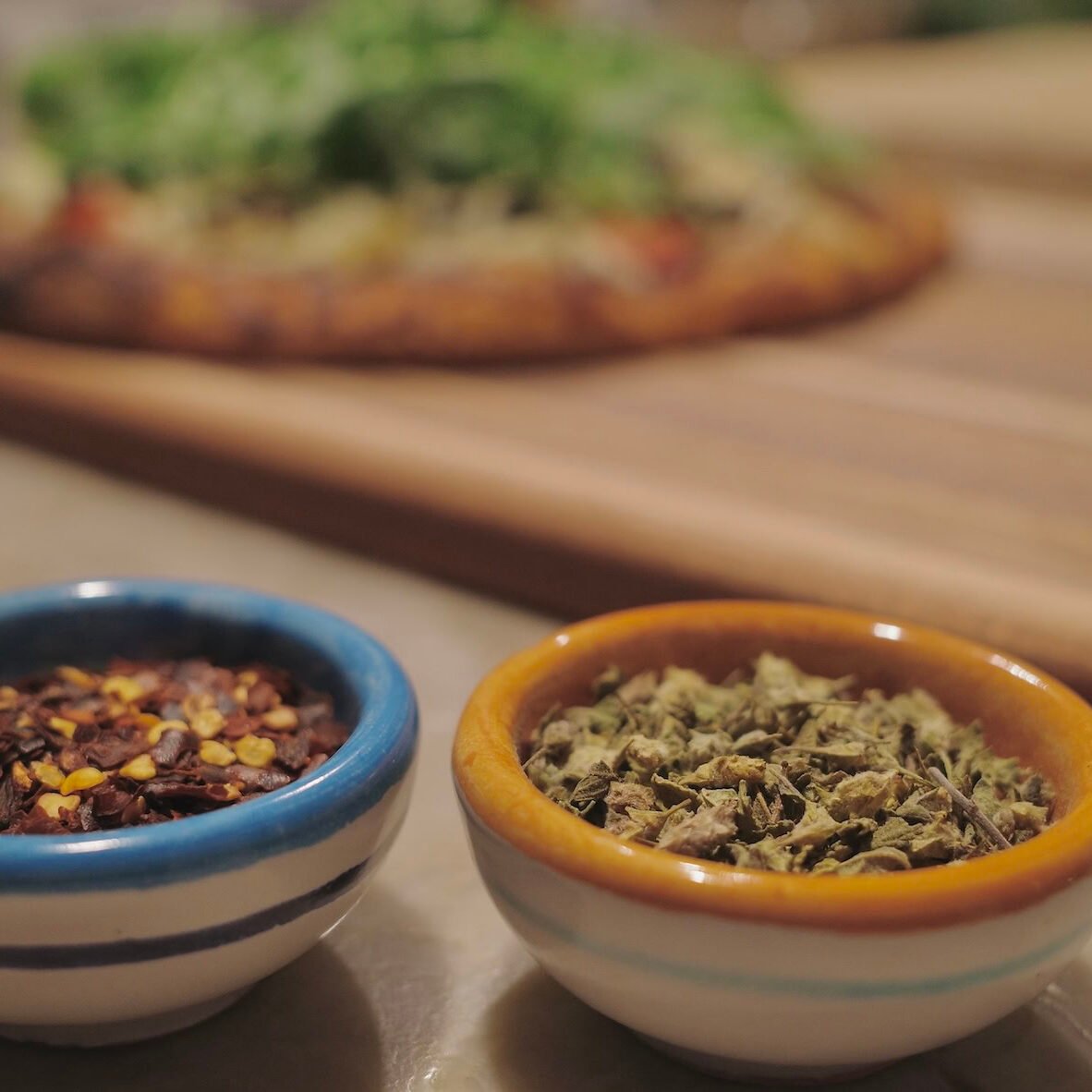 Dried oregano and crushed red pepper in small bowls with a pizza in the background