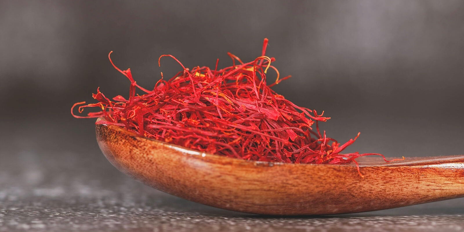 A heap of deep red saffron threads (Crocus sativus) piled in a polished wooden spoon, close-up showing the fine, wiry, slightly trumpet-tipped strands