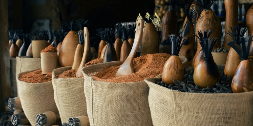 A traditional spice market stall with hessian sacks full of deep red ground spice and dark whole spice, surrounded by carved wooden gourd vessels with black tasseled tops and wooden scoops resting in the powder
