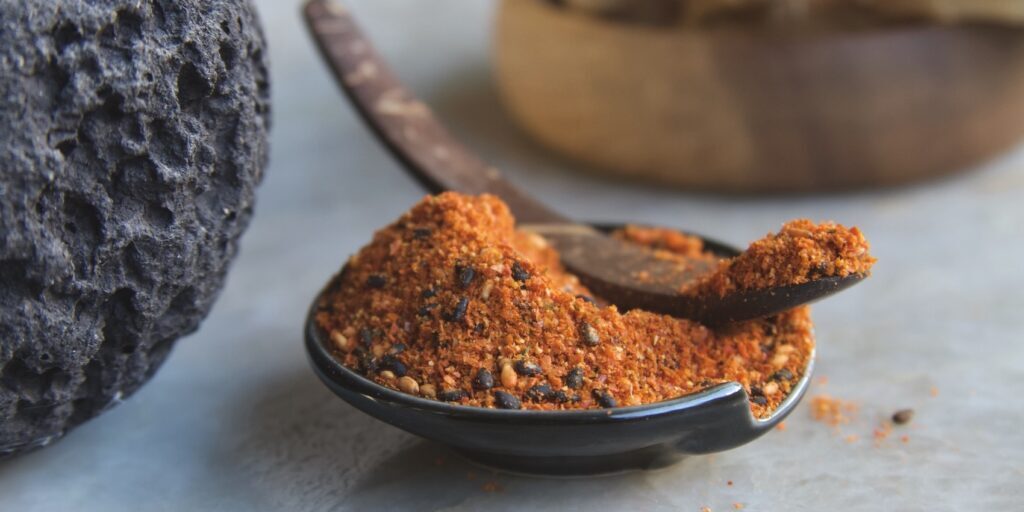 Shichimi togarashi Japanese seven-spice blend mounded in a black ceramic dish with a curved wooden spoon showing vivid orange-red color and visible black sesame seeds